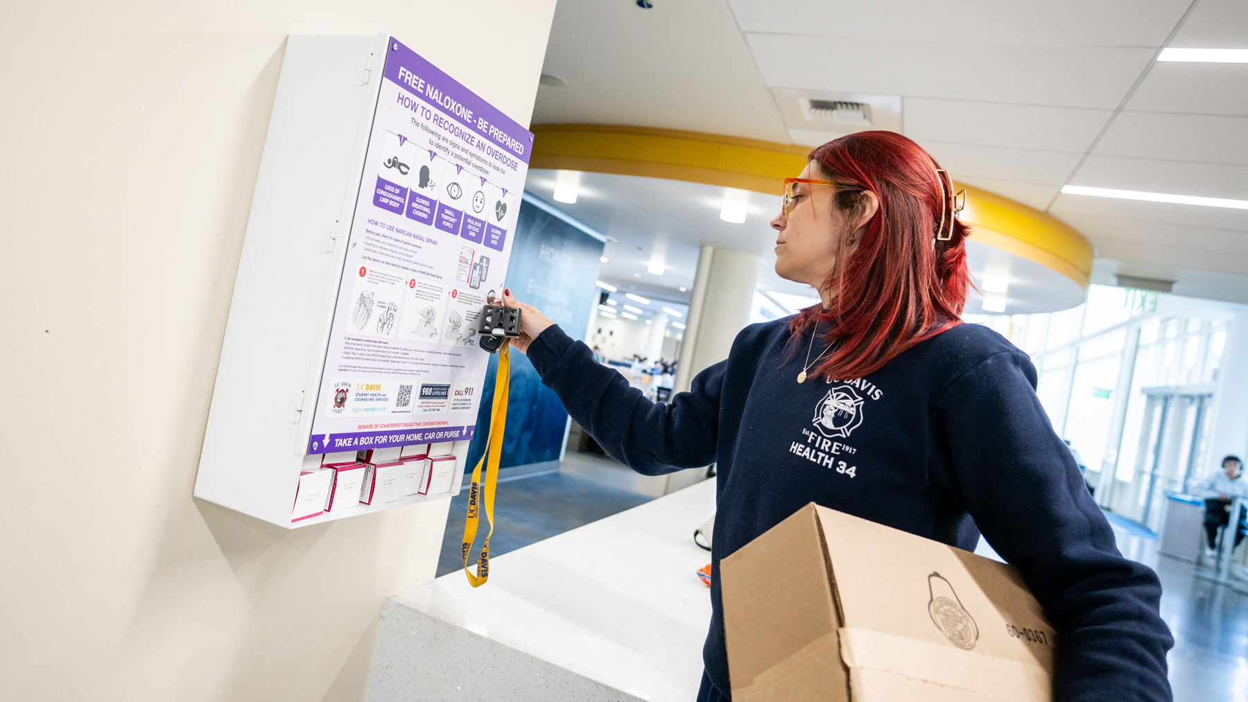 An EMT in a UC Davis shirt with a box of supplies, unlocks the Narcan cabinet in the Memorial Union